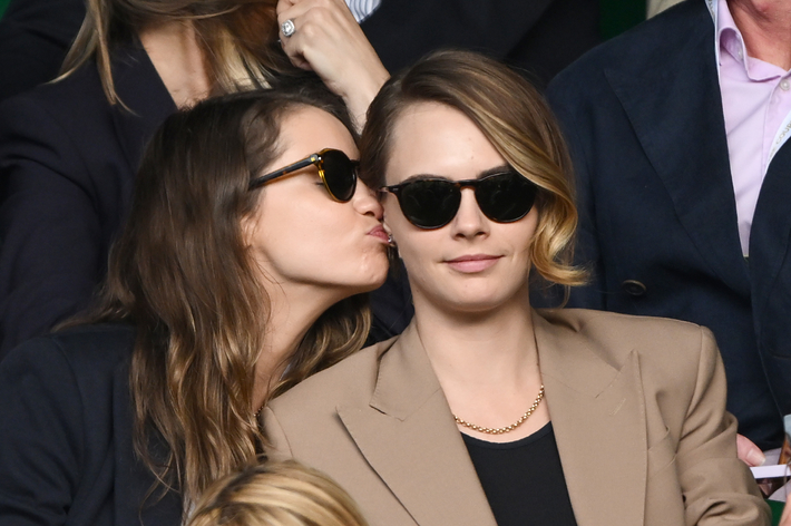 Cara Delevingne and Ashley Benson at an event. Cara in a beige blazer and black top, Ashley in a black jacket, both wearing sunglasses. Ashley kisses Cara's cheek