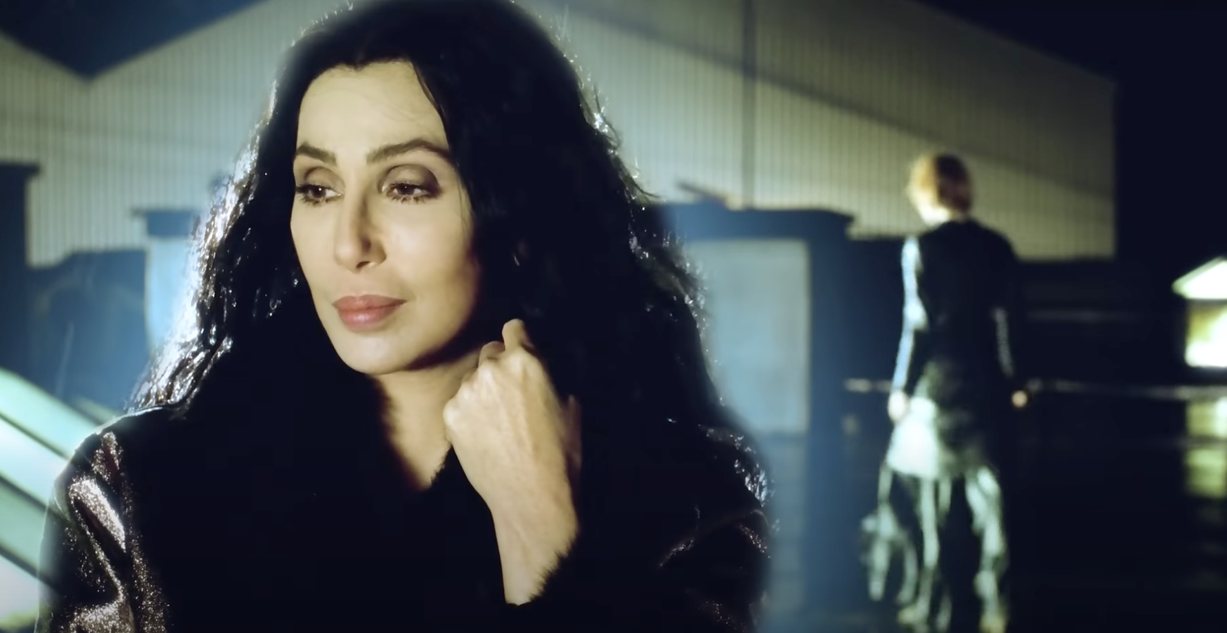 Cher in a reflective pose with a building backdrop; another person in a modern outfit is seen from behind in the distance