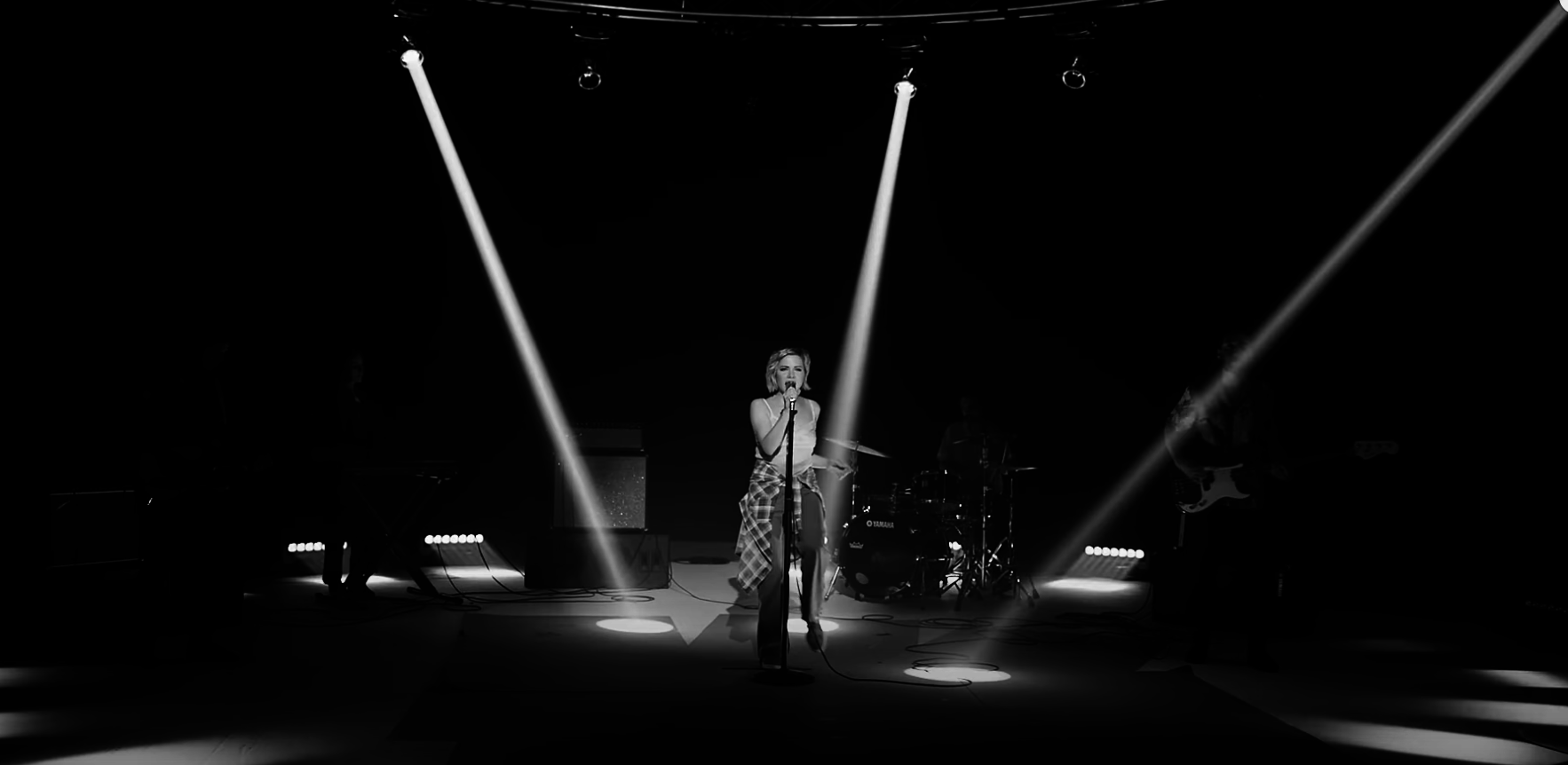 Black and white image of a singer performing on stage with a microphone, illuminated by spotlights. Band members and instruments are in the background. No names provided