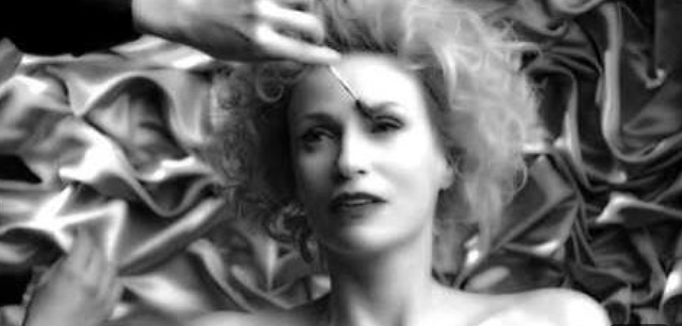 Gillian Anderson in a black-and-white photo shoot, being styled by a hand using a makeup brush against a textured backdrop