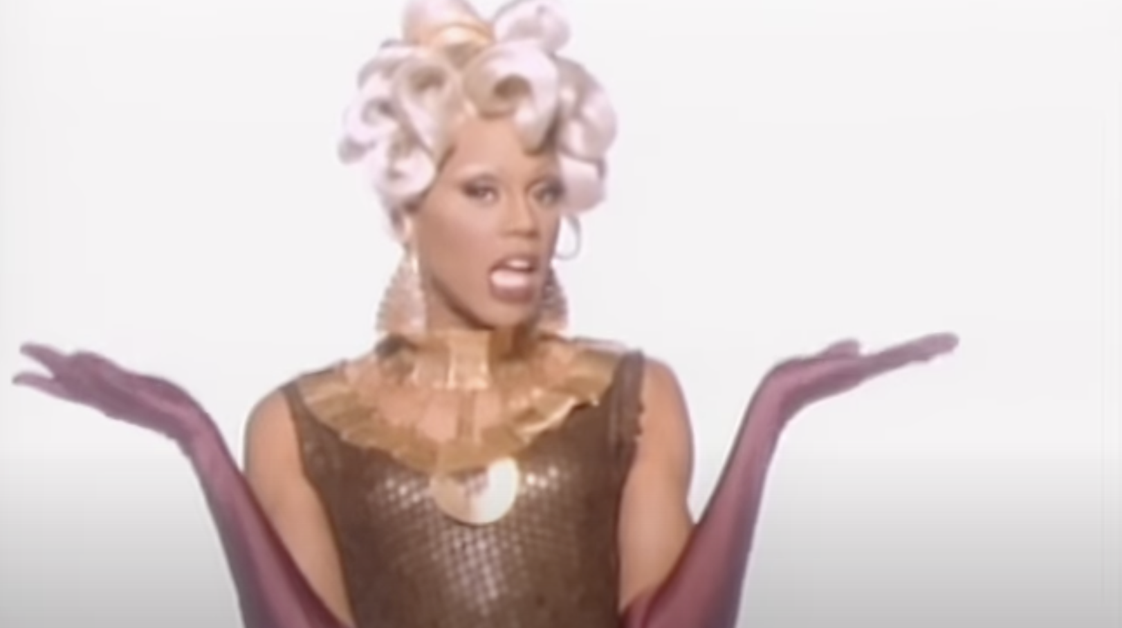 RuPaul poses with an expressive gesture, wearing a sequined outfit, gold accessories, and curly hair styled upwards