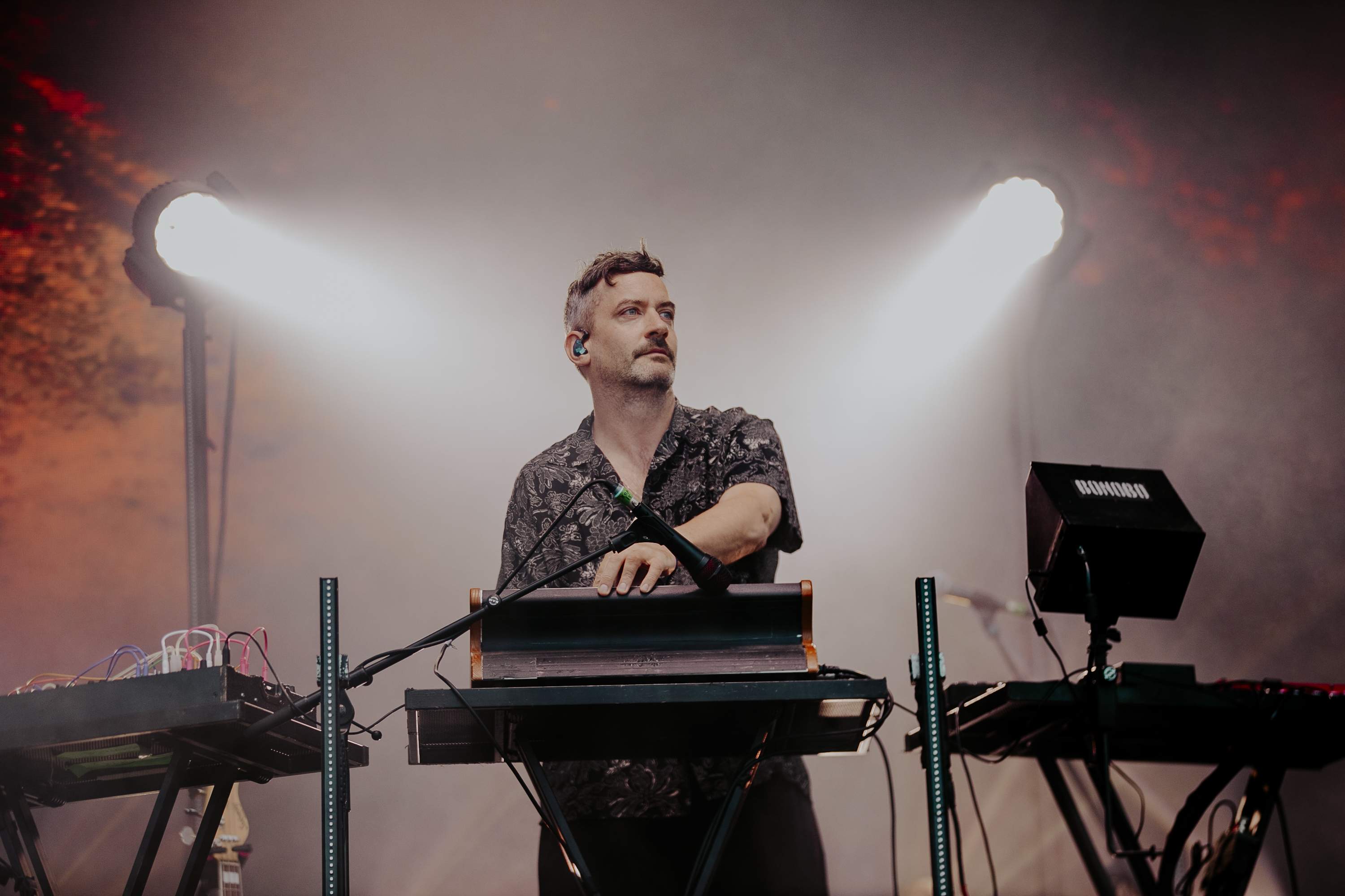 A person stands behind electronic music equipment on stage, with lights shining behind them. The person is looking to the side and wearing a patterned shirt