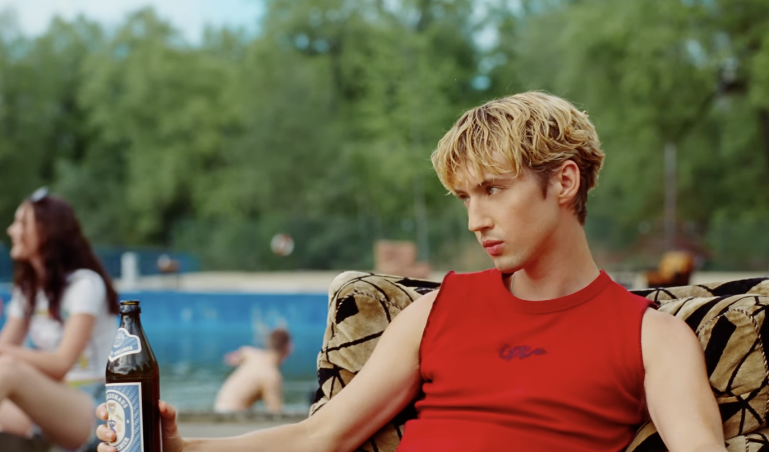 Troye Sivan in a sleeveless top sits on a patterned chair holding a beer bottle at an outdoor pool area. Another person is blurred in the background