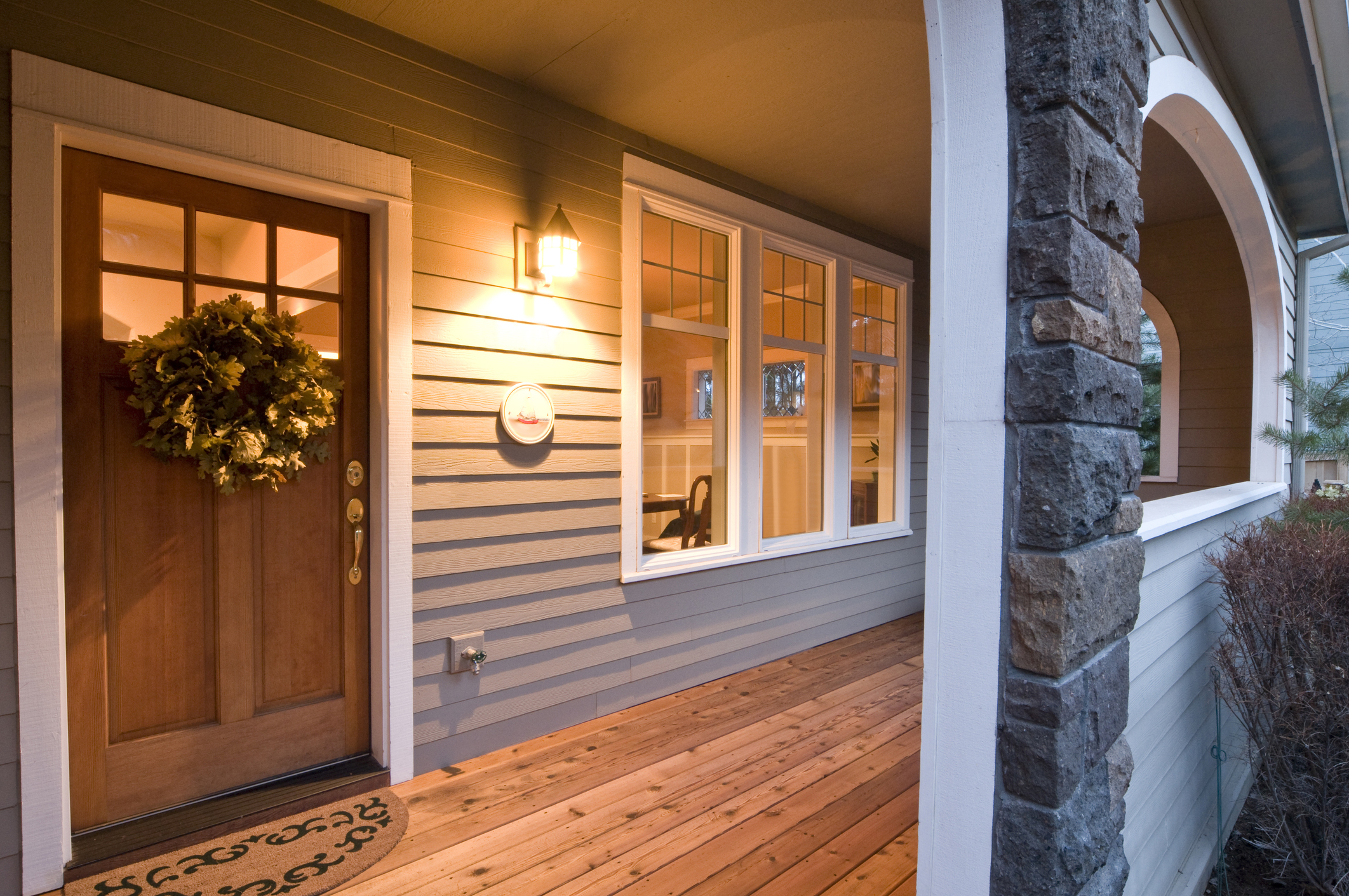 Front porch of a house with wooden flooring, a wreath on the door, and an arched stone column on the right side