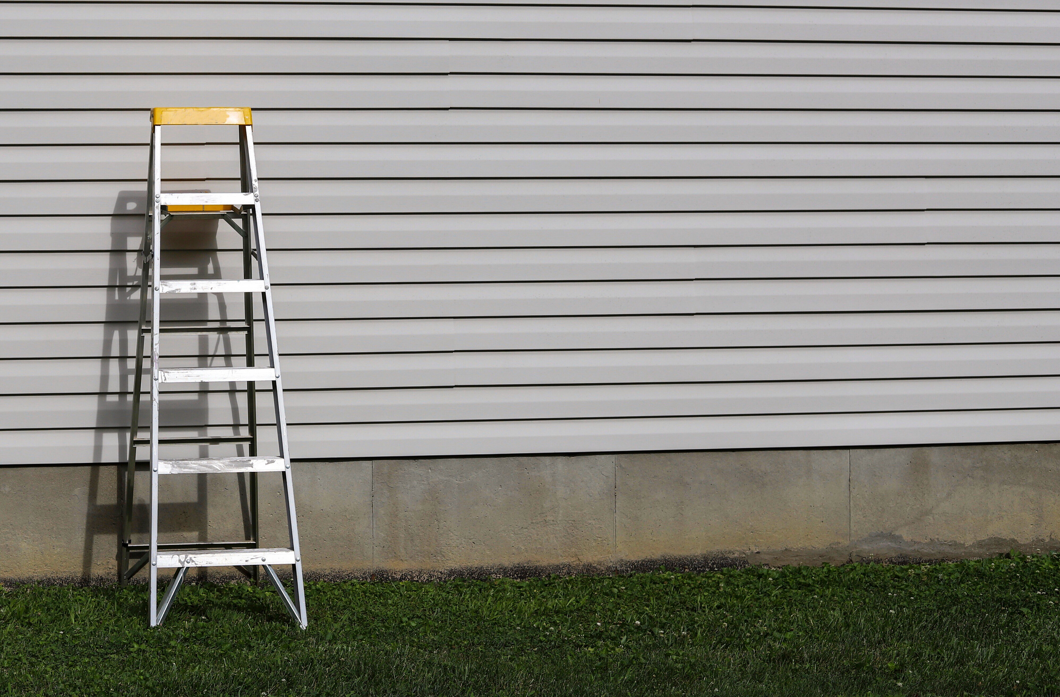 A ladder is leaned against the side of a house with horizontal siding, standing on a grassy area next to a concrete foundation