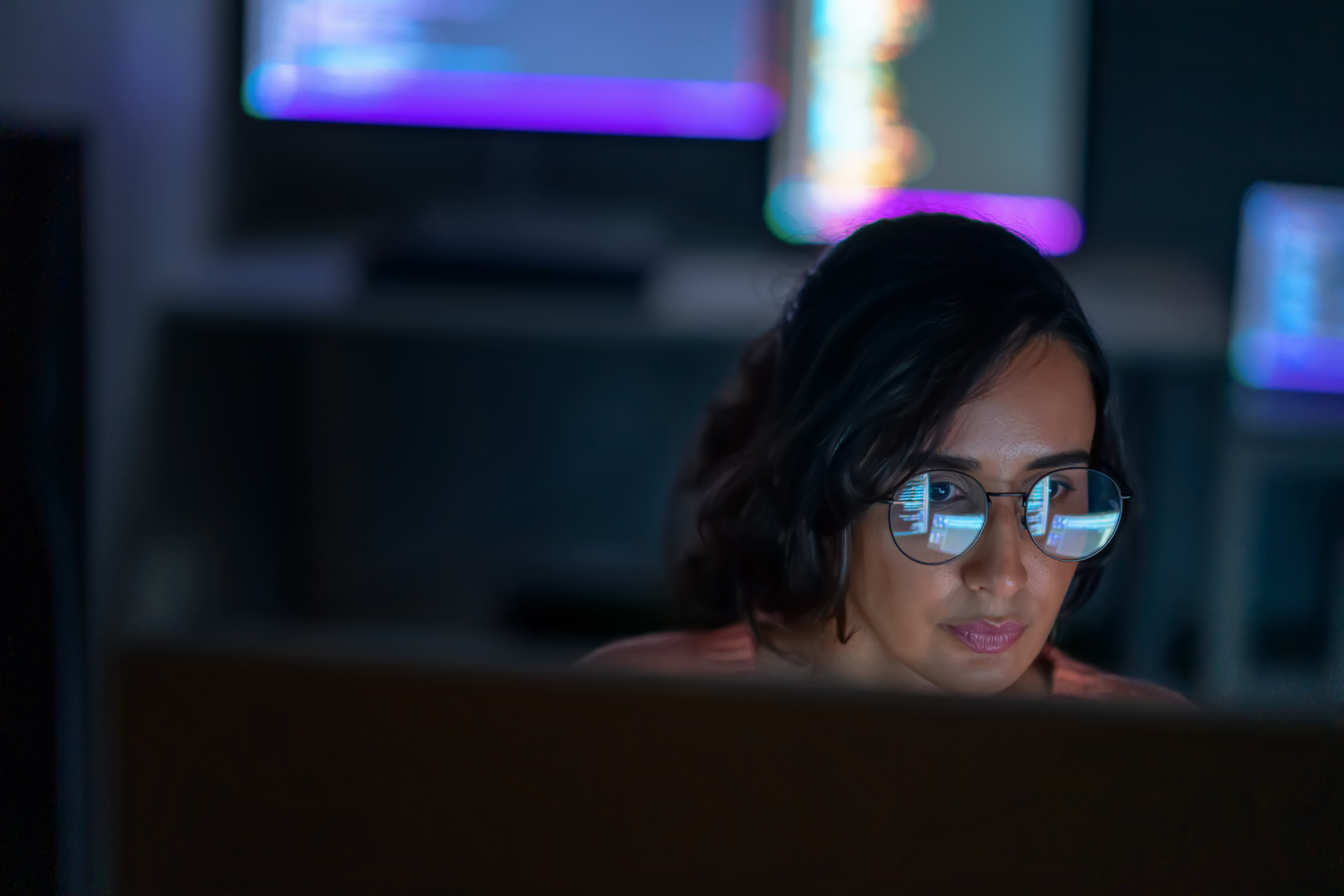 A person wearing glasses works on a computer, with code reflected in their glasses and multiple computer screens in the background