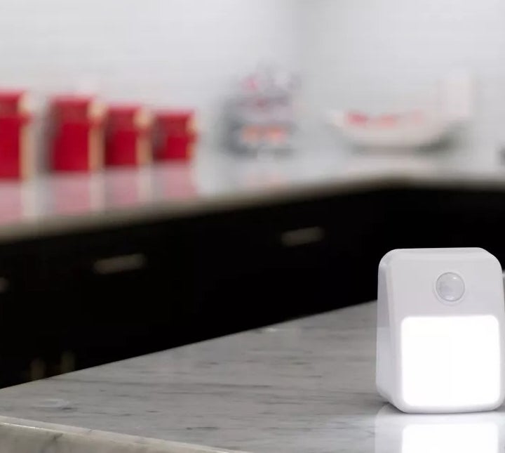 A small white motion-sensor night light sits on a marble kitchen countertop with storage bins and a bowl in the blurred background