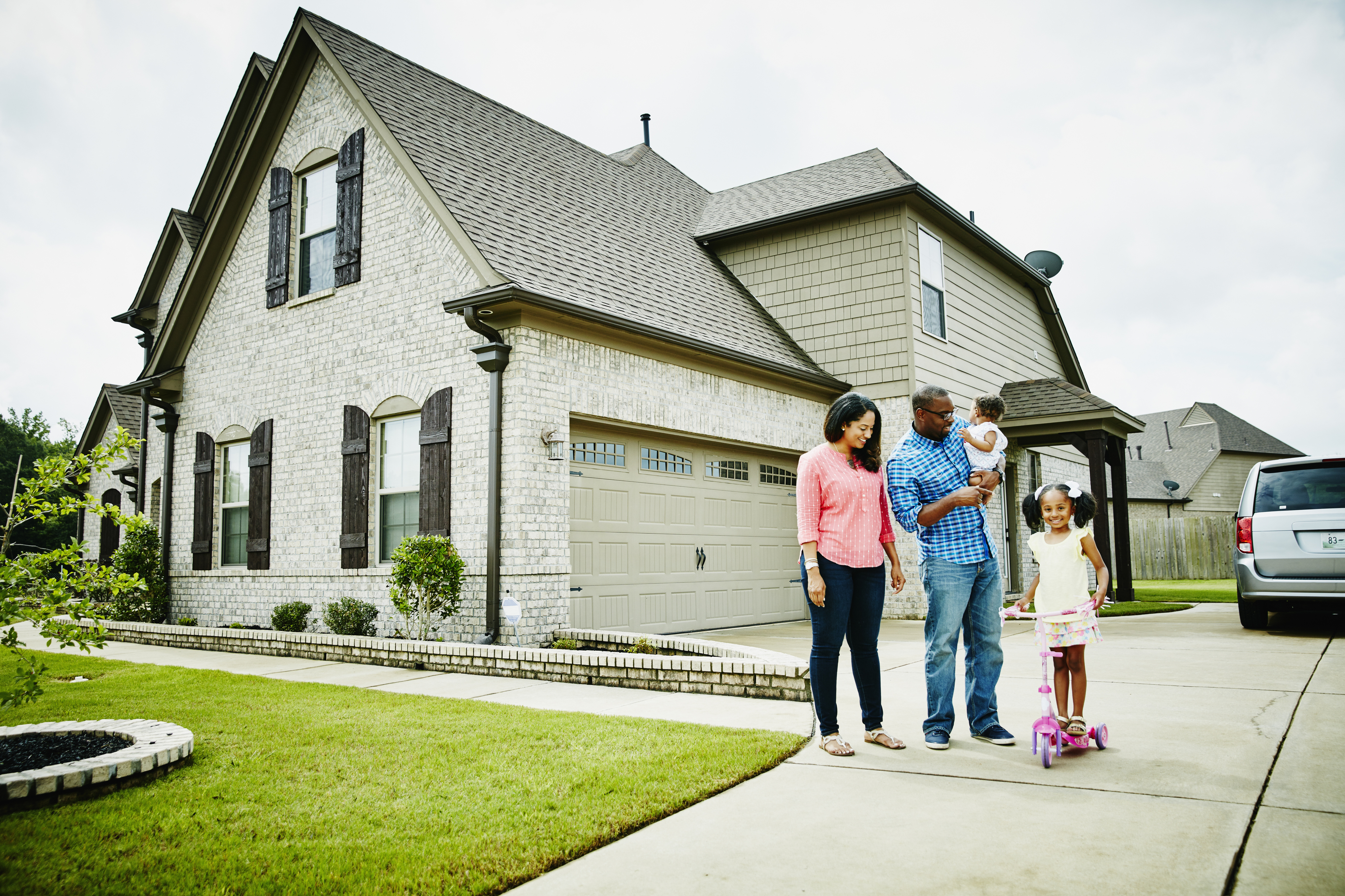 A family of four enjoys time outside their suburban home. The mother and father watch as their two children, one on a scooter, play on the driveway