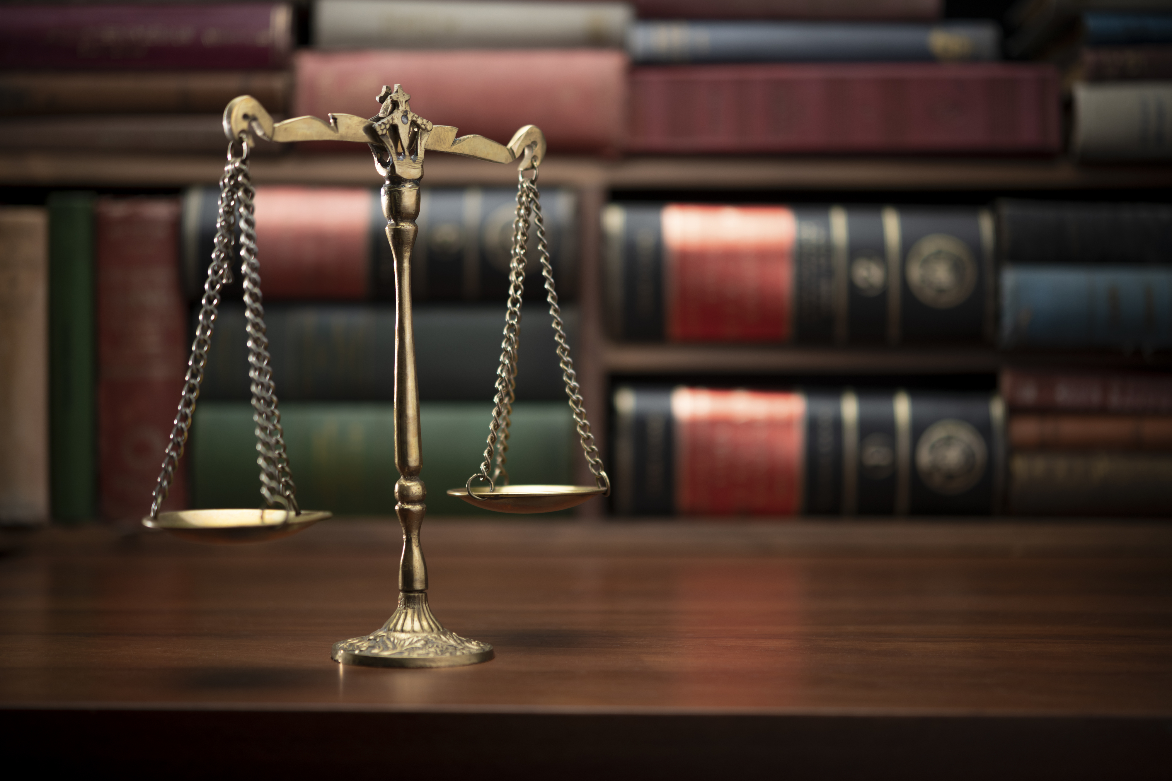 A balanced scale sits on a wooden desk in front of a row of library books, symbolizing justice and fairness