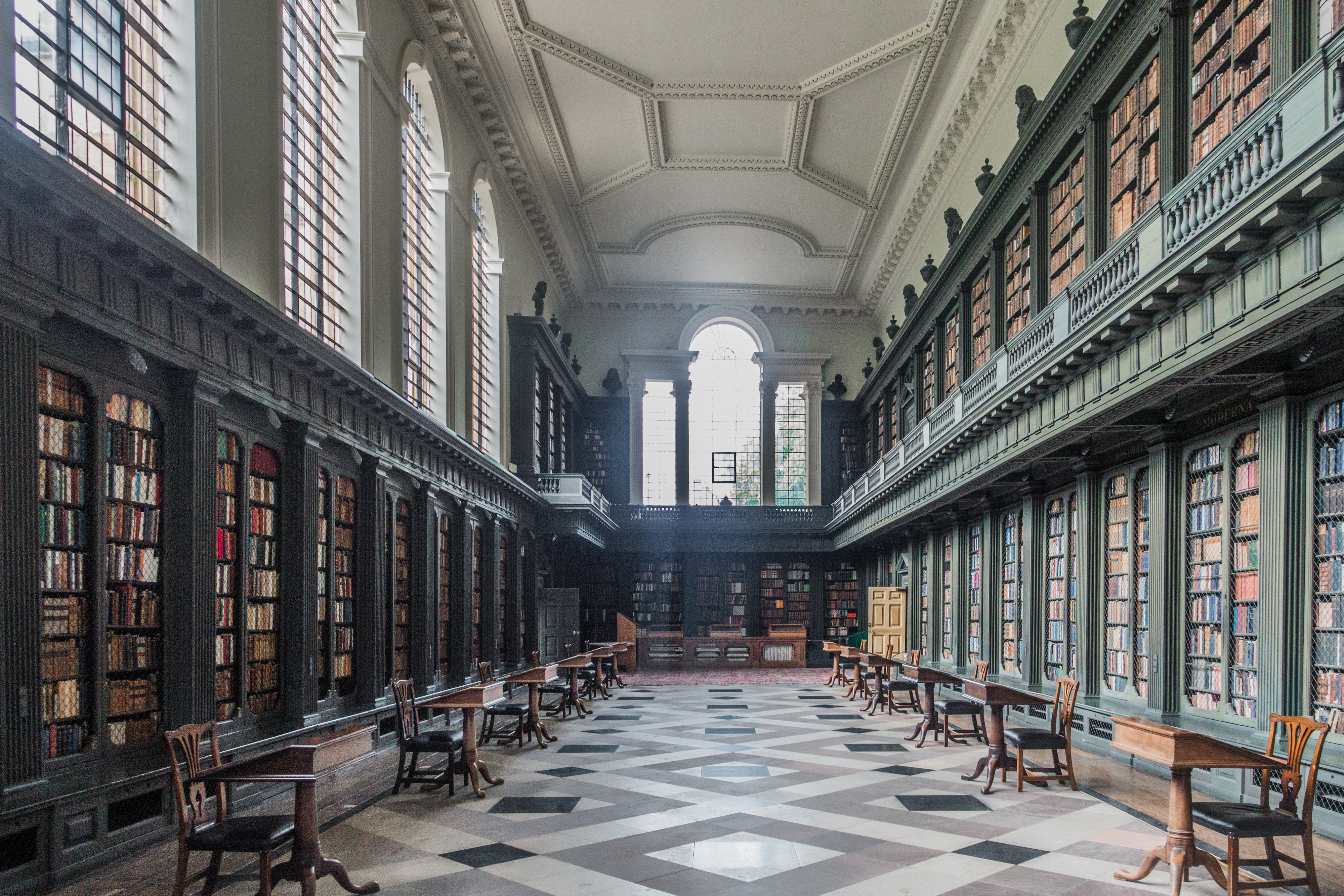 Interior view of a large, historic library with tall bookshelves lining the walls, long wooden tables, and chairs organized neatly on a patterned floor