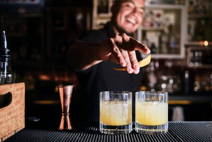 A bartender smiles as he garnishes two cocktails with lemon peels in a dimly lit bar