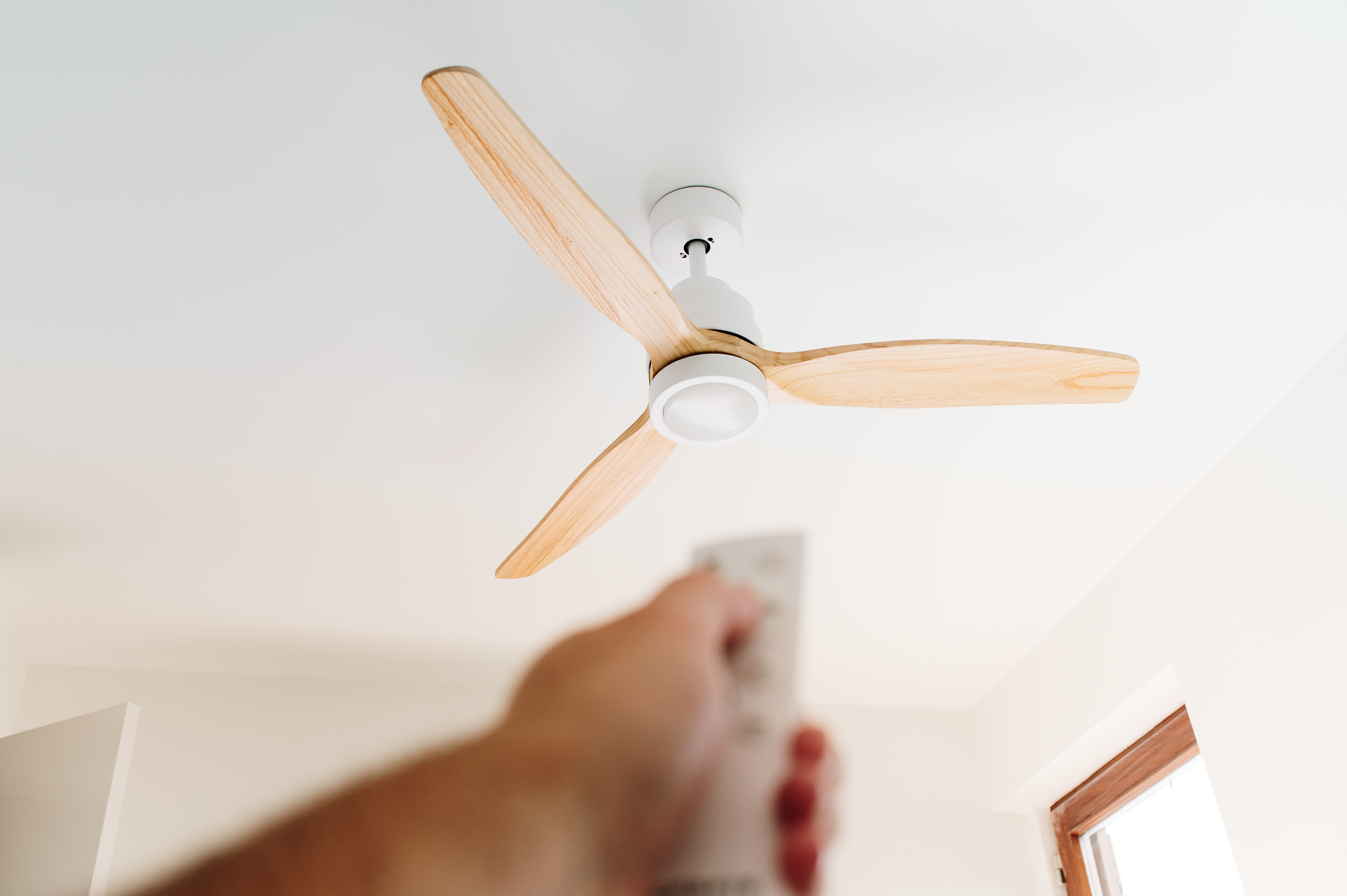 A hand holds a remote control, pointing it at a ceiling fan with three wooden blades