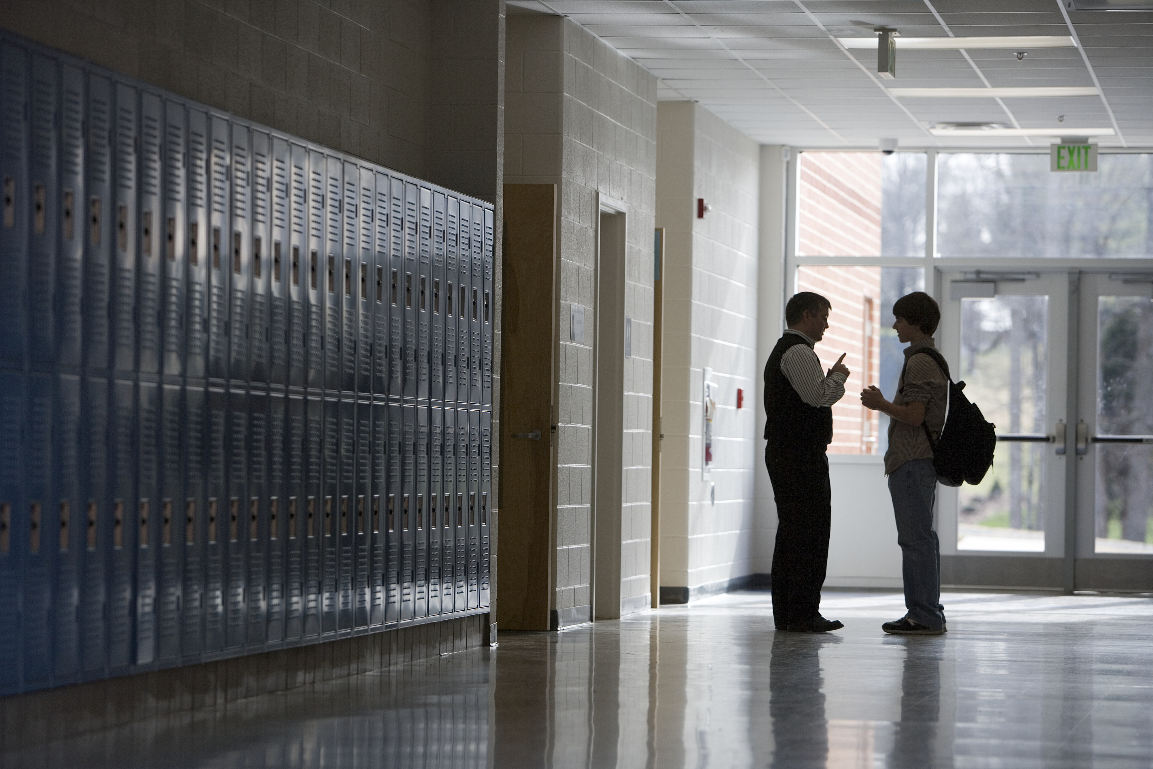 A teacher talks to a student in a quiet school hallway lined with lockers. The teacher gestures with their hand as they engage in conversation