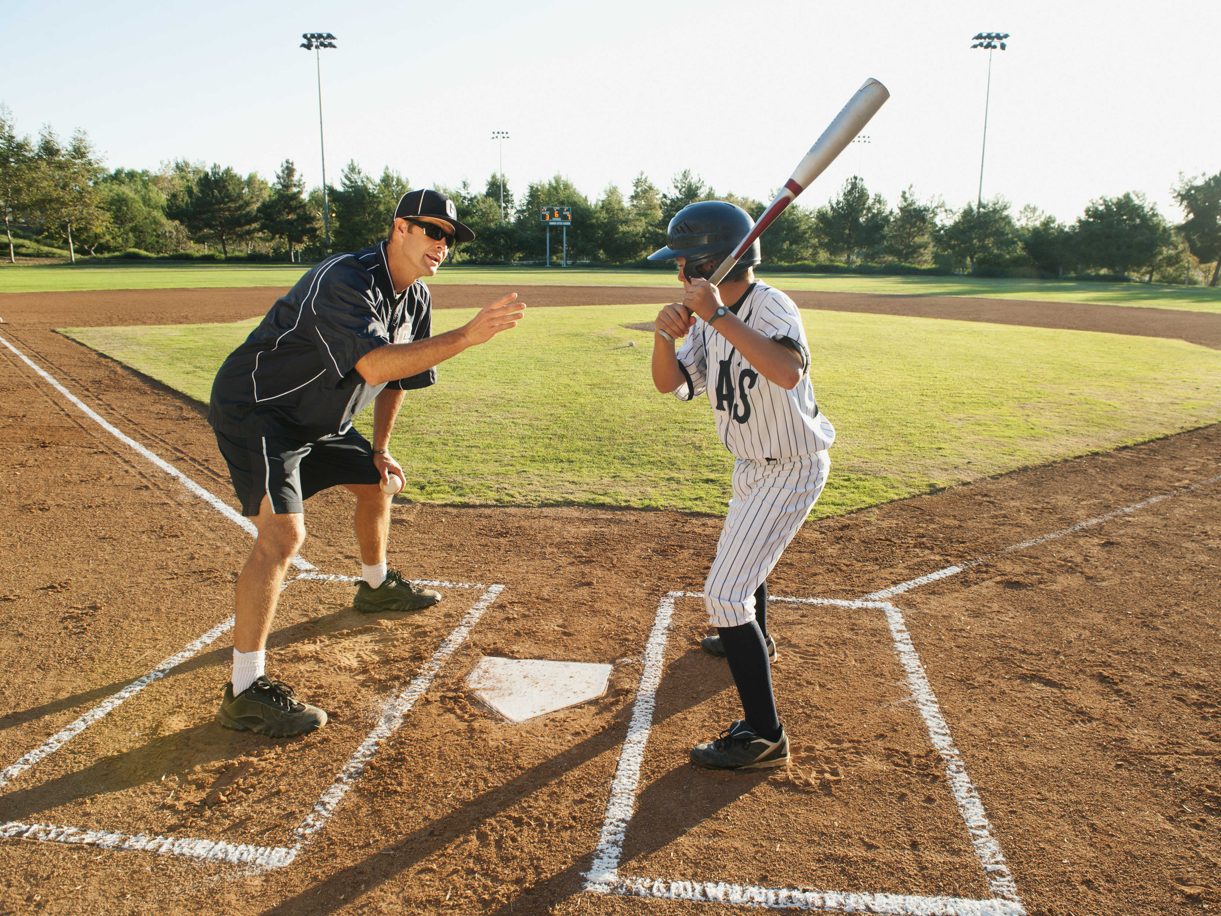 A coach instructs a young baseball player at home plate on a baseball field. Both wear sports attire, with the player in a helmet and holding a bat