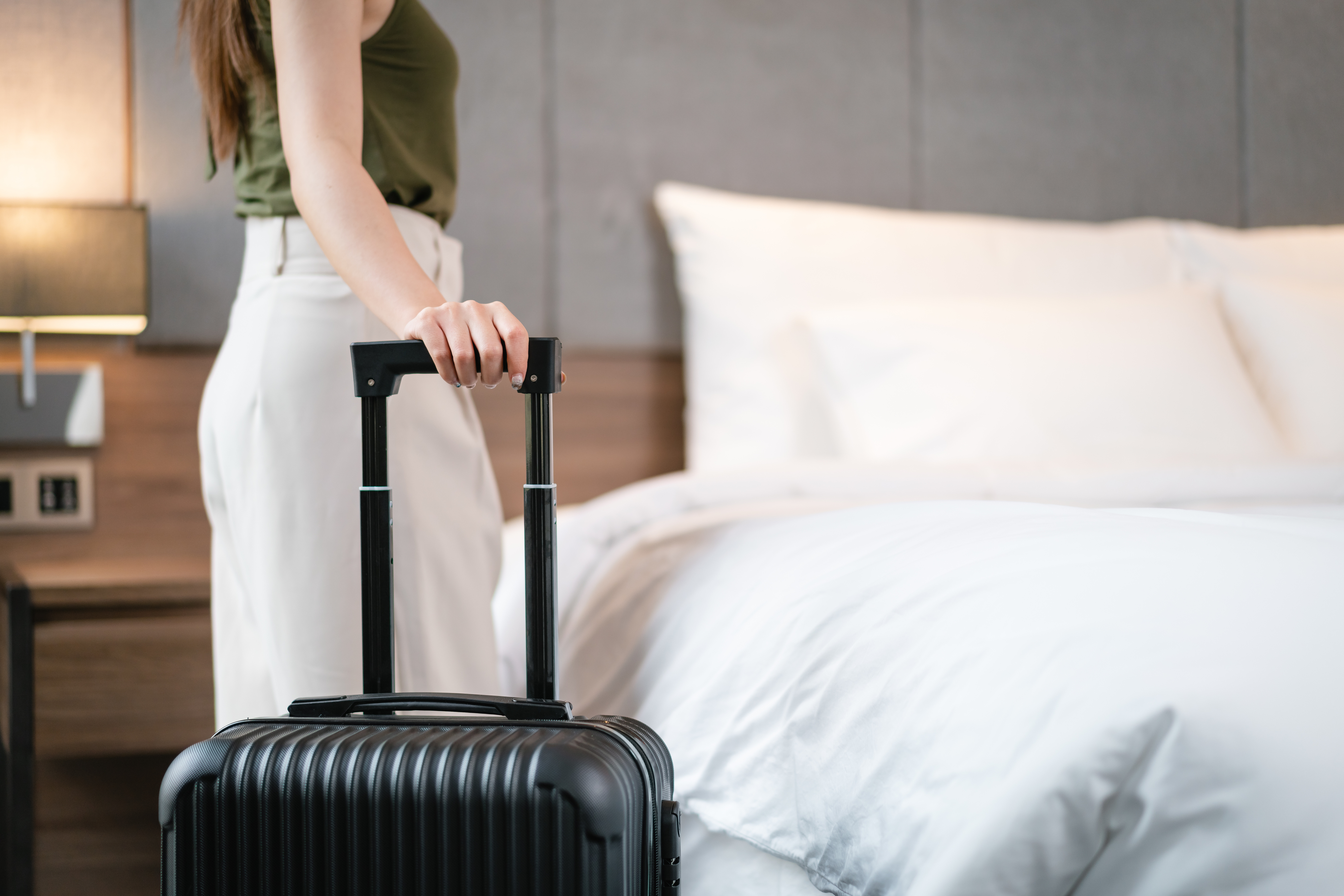 A person standing beside a bed, holding the handle of a roller suitcase in a bedroom. Only the person’s arm and part of their torso are visible