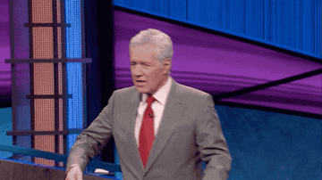 Alex Trebek in a suit and red tie, animatedly walking on the set of &quot;Jeopardy!&quot; with a background featuring the show&#x27;s signature colored lights and podiums