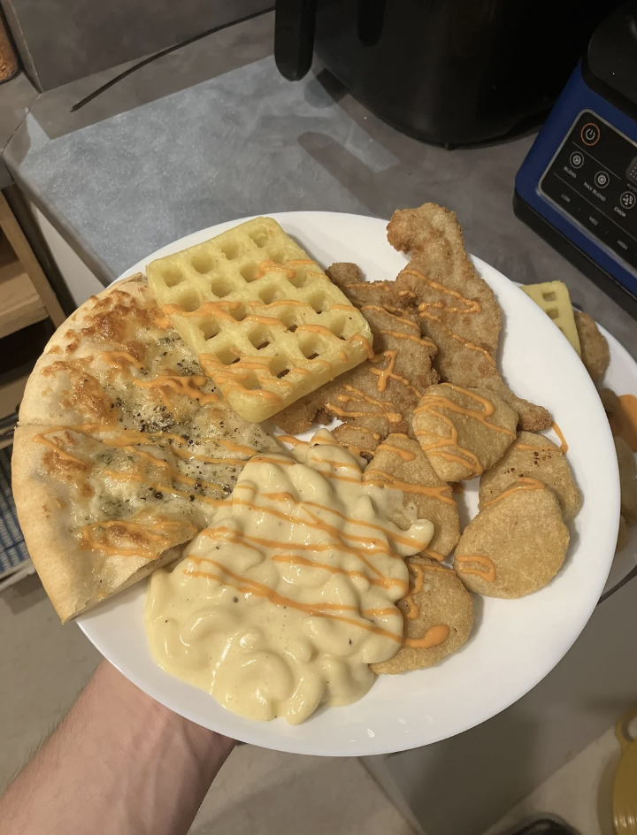 A plate of various foods, including waffle, chicken nuggets, slices of pizza, cooked macaroni and cheese, and chicken tenders, with a drizzle of sauce