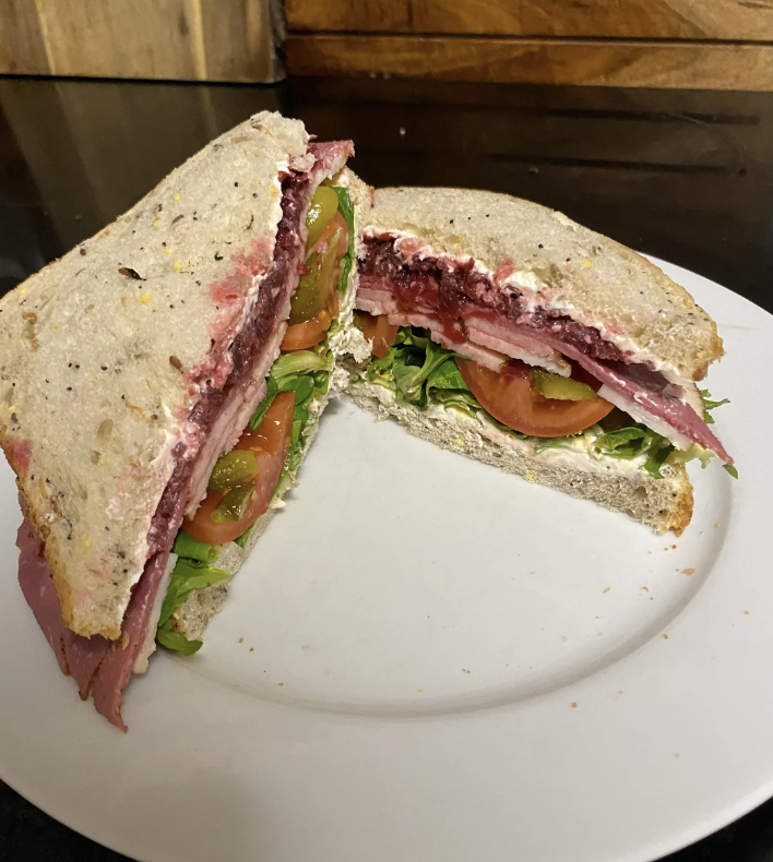 A sandwich on a plate, filled with layers of meat, fresh greens, tomato slices, and a spread