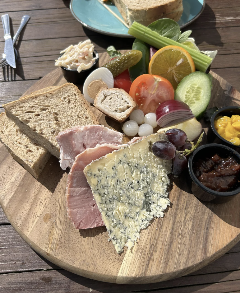 A wooden platter featuring various cheeses, slices of bread, ham, grapes, pickles, cucumber, tomato, and small dishes with pickles and chutney, on a wooden table