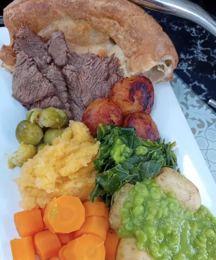 A plate featuring roast beef, Yorkshire pudding, roast potatoes, mashed turnips, brussels sprouts, carrots, kale, and potatoes topped with mushy peas