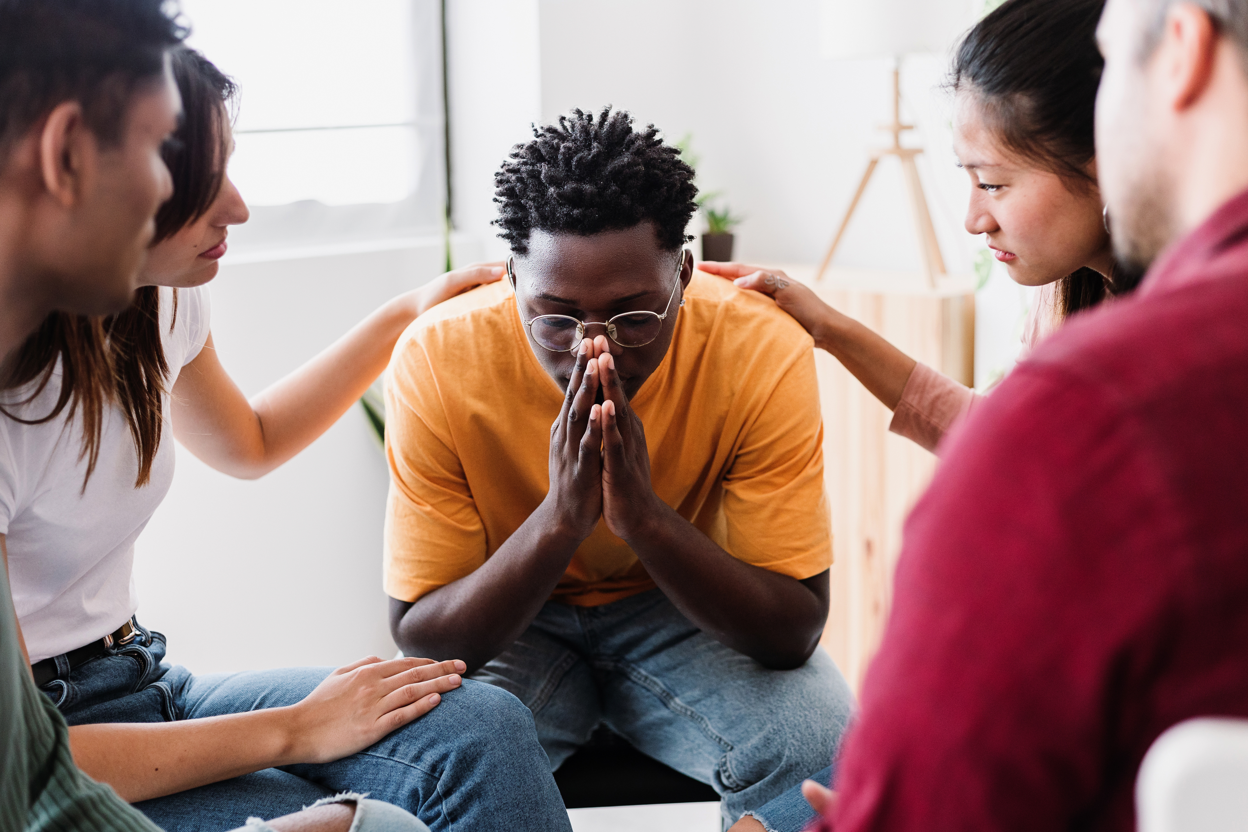 A group of five people sit in a circle, offering comfort and support to a distressed young man in the center who clasps his hands in front of his face