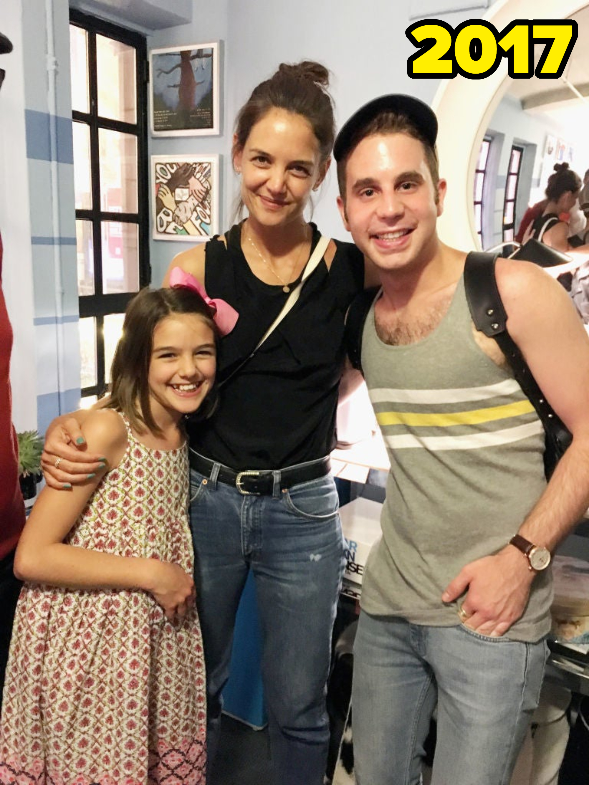 Katie Holmes, Suri Cruise, and Ben Platt are smiling together indoors. Katie and Suri are casually dressed; Ben is in a tank top and cap