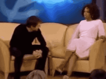 Tom Cruise energetically jumps on a couch next to Oprah Winfrey during an interview on her talk show. Oprah looks surprised and laughs