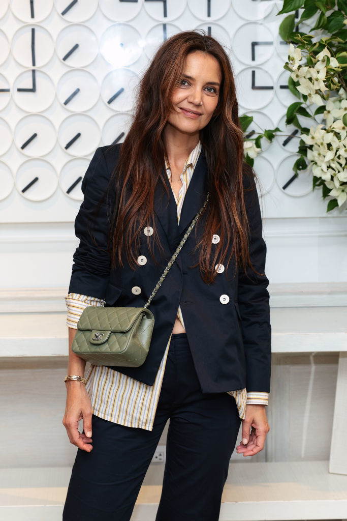 Katie Holmes is wearing a black blazer with white buttons, a striped shirt underneath, and carrying a quilted handbag. She poses in front of a decorative wall