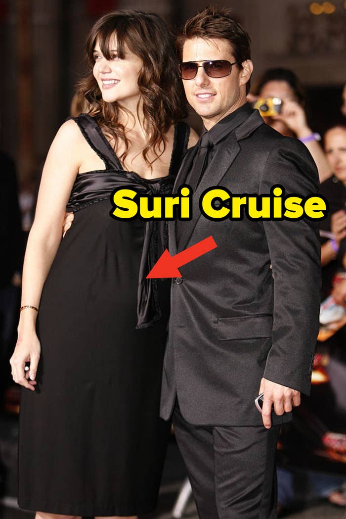 Katie Holmes and Tom Cruise posing together on a red carpet. Katie is in a sleeveless black dress with a bow, and Tom is wearing a black suit with sunglasses