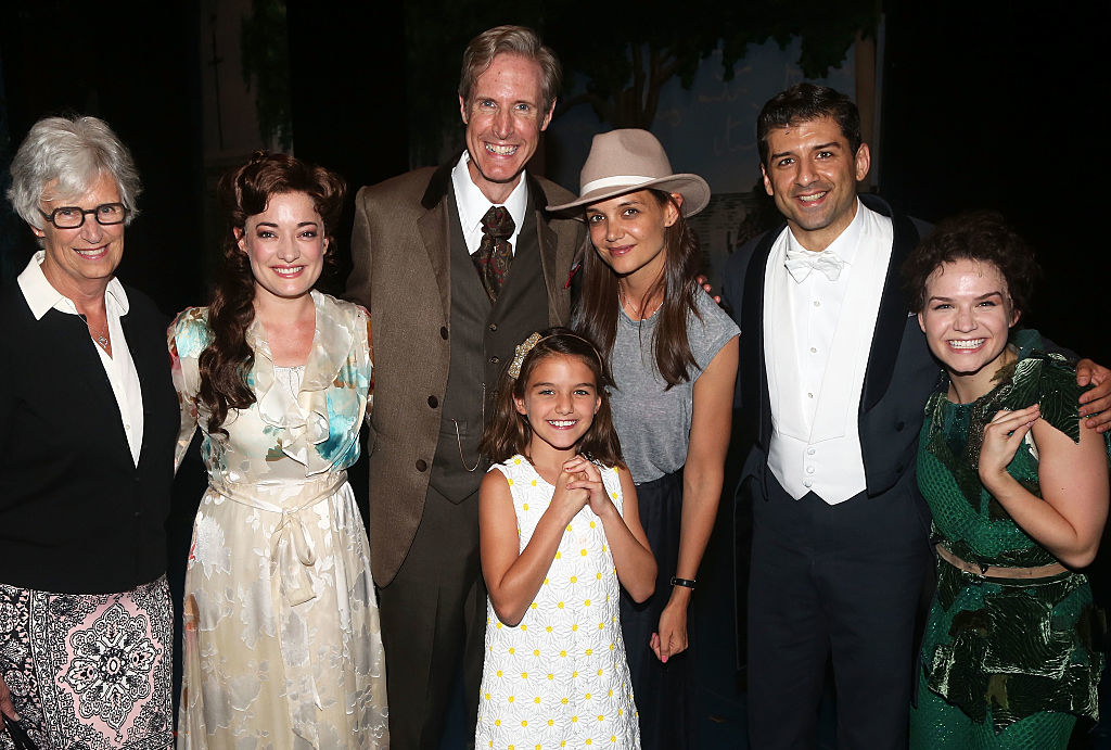Group photo of a Broadway cast with five adults (one wearing a broad hat) and two girls posing together and smiling