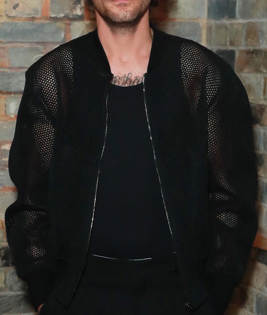 Louis Tomlinson stands against a brick wall, wearing a black mesh jacket, black shirt and black pants