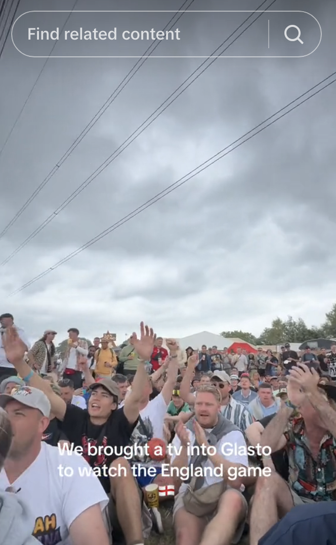A crowd of people sitting and raising their hands while watching a TV at Glastonbury Festival. Text reads, &quot;We brought a TV into Glasto to watch the England game.&quot;