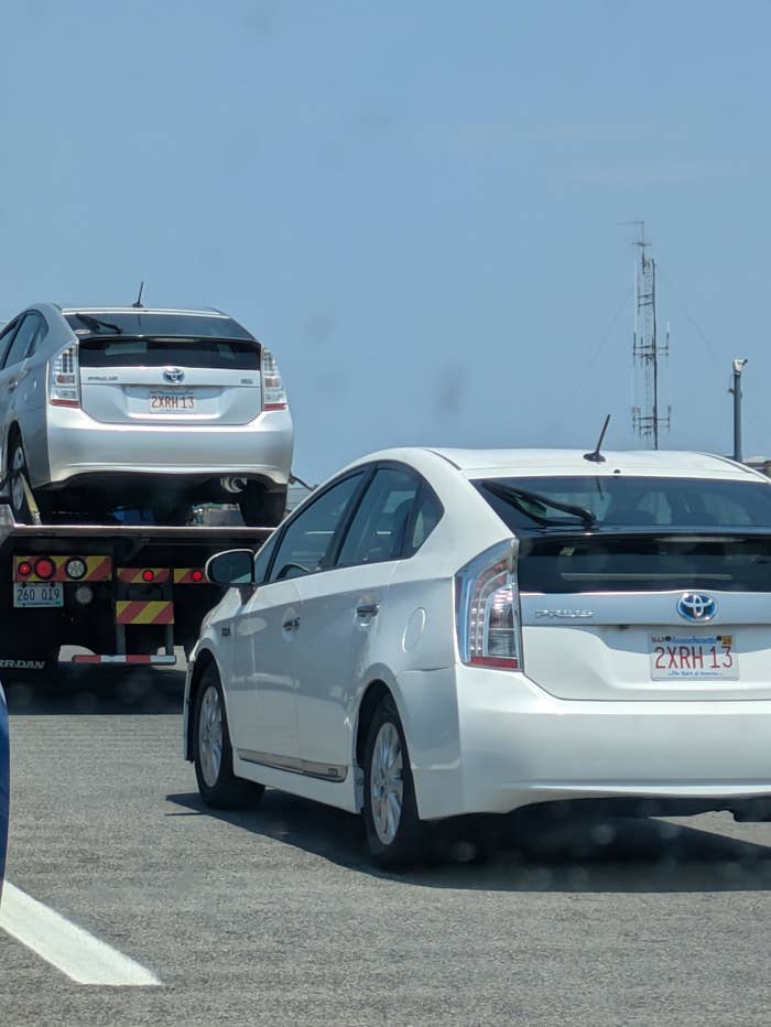 Two Toyota Prius cars on a highway, one of them loaded onto a tow truck. Both cars have the exact same license plates