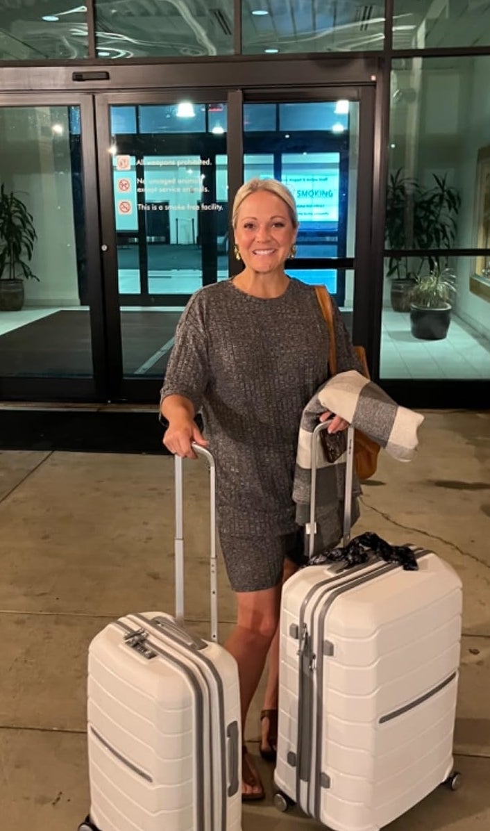 A reviewer in stylish business attire is standing outside, holding two white rolling suitcases, ready for travel