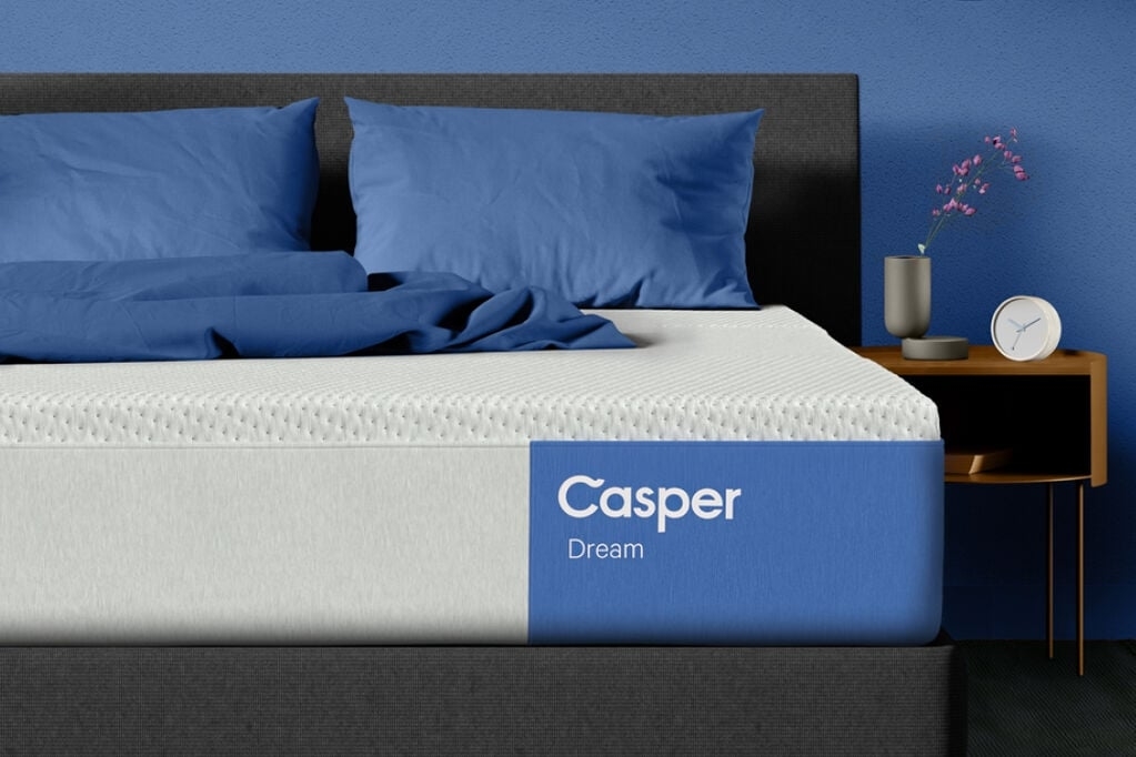 Casper Dream mattress on a modern bed with blue pillows and bedding, a small nightstand with a vase holding a pink flower, a clock, and a candle