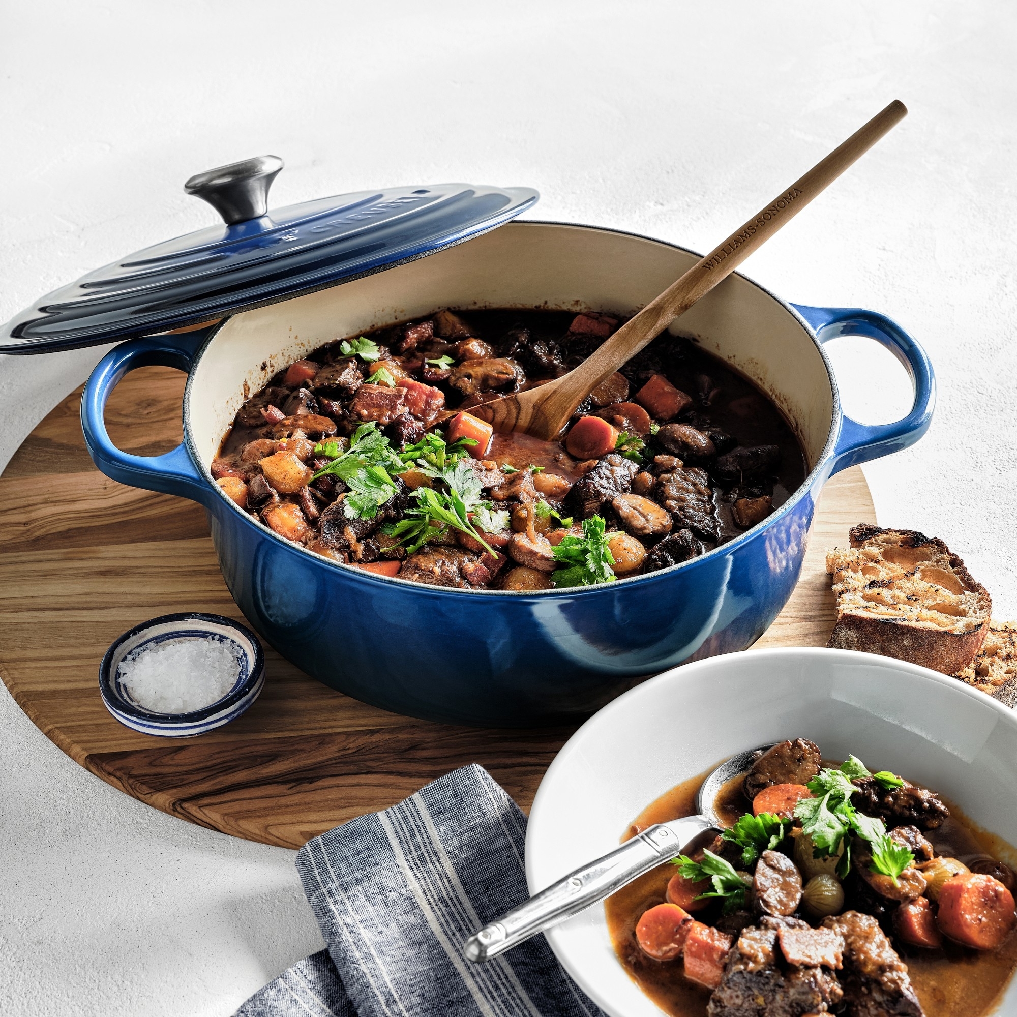 A blue Dutch oven filled with hearty beef stew sits on a wooden board, with a serving spoon inside. A bowl of stew and a striped napkin are next to it