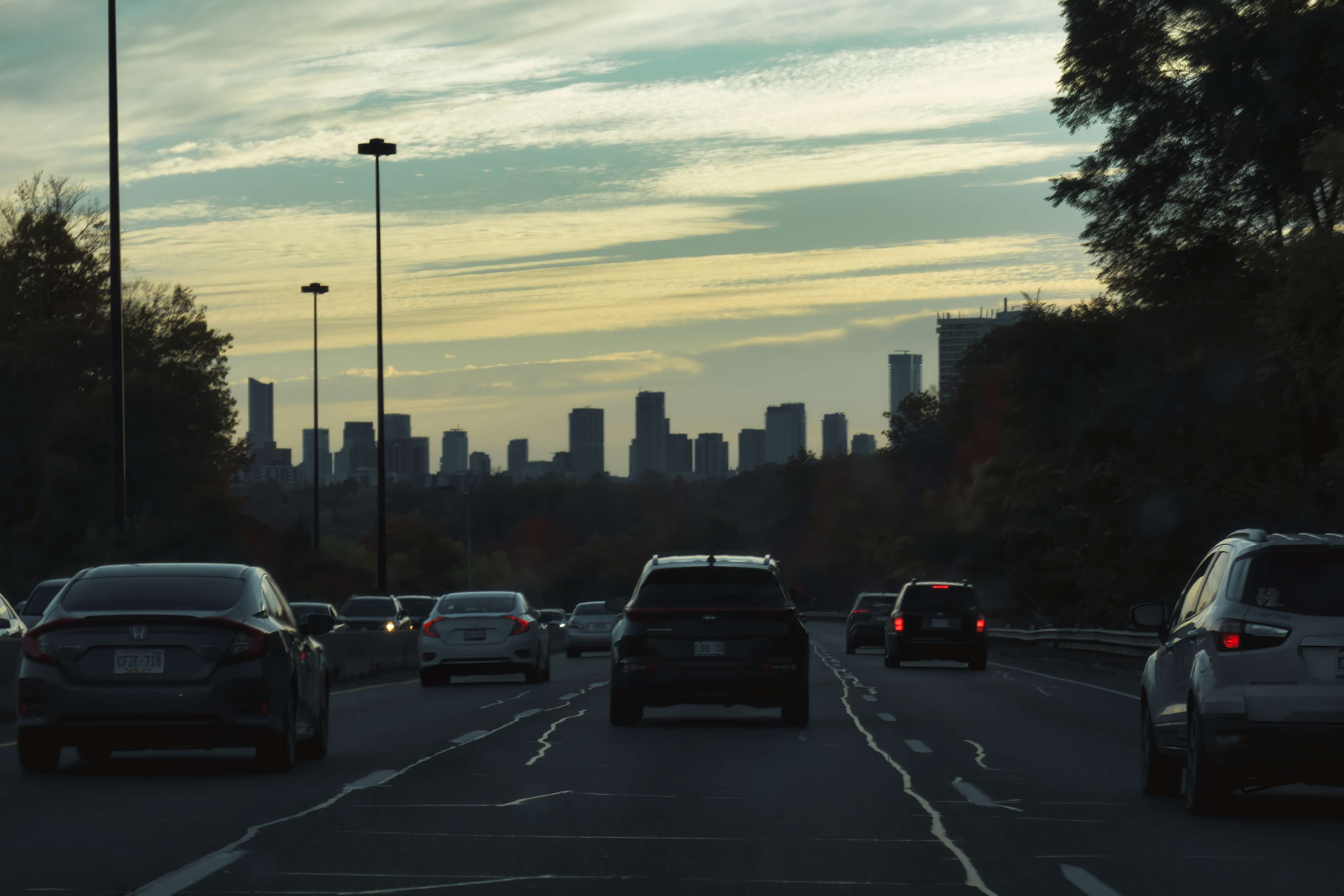 Cars driving on a highway towards a city skyline at dusk. The sky is streaked with clouds