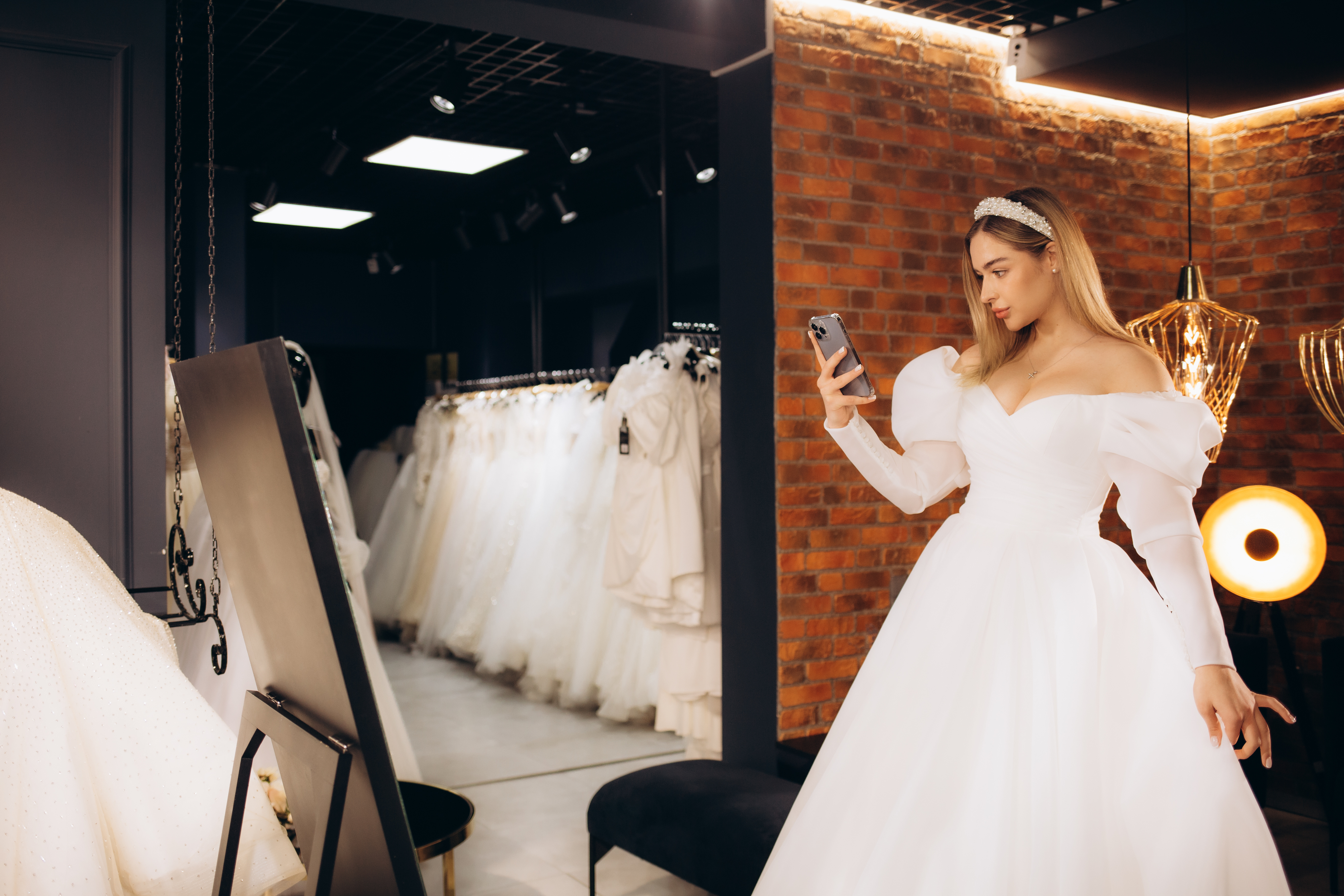 A woman in an off-the-shoulder wedding dress takes a selfie in a bridal boutique
