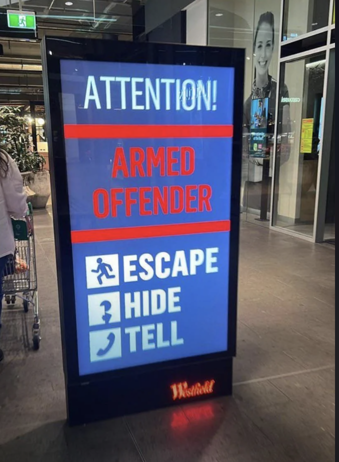A digital sign in a shopping center displays a warning about an armed offender with instructions to escape, hide, and tell
