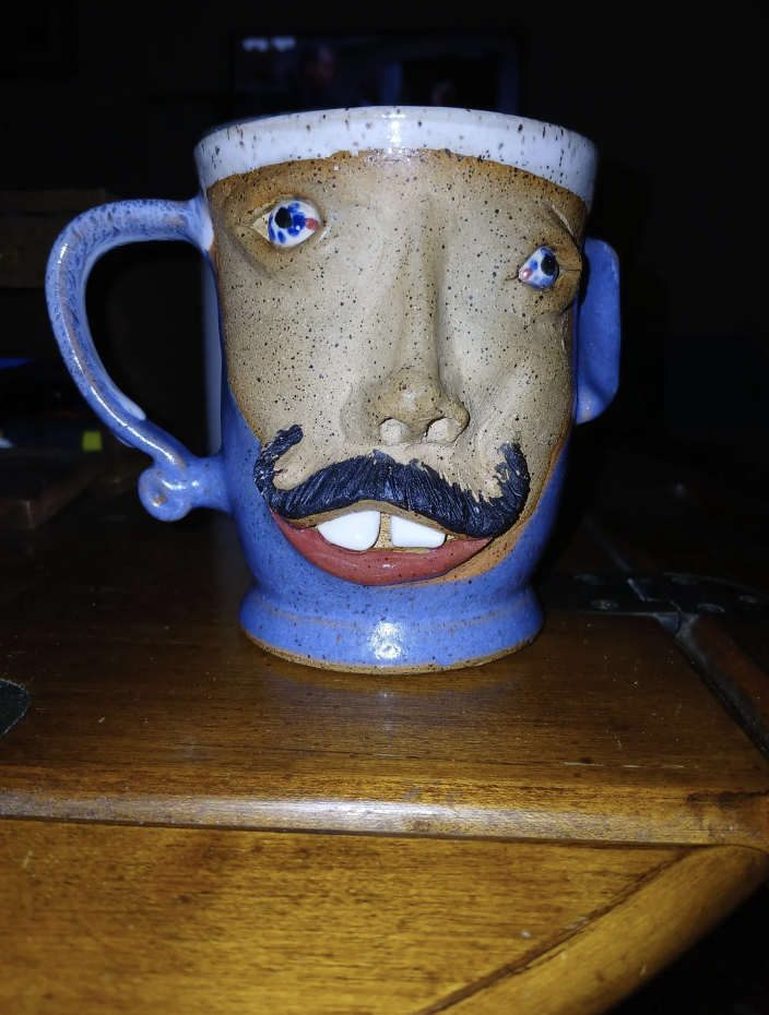 A handcrafted ceramic mug with a face design, featuring a mustache, nose, and protruding teeth