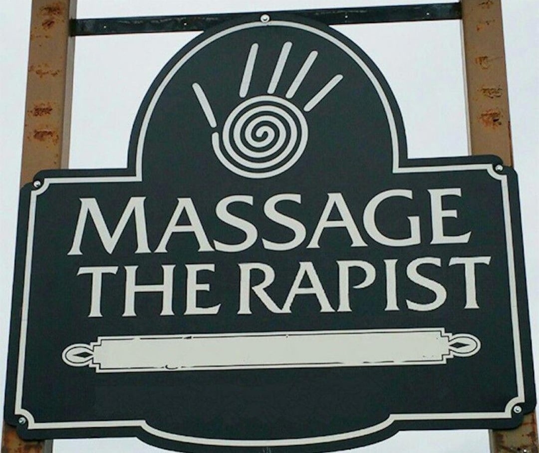 An outdoor sign that reads &quot;Massage the rapist&quot; with a decorative hand symbol at the top; there&#x27;s some very unfortunate spacing going on (it&#x27;s supposed to read Massage Therapist)