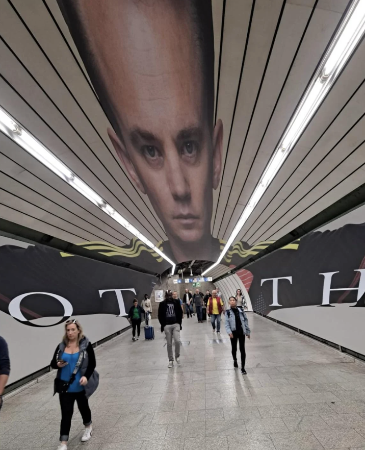 Subway station with a large, distorted face poster on the ceiling while people walk in various directions below