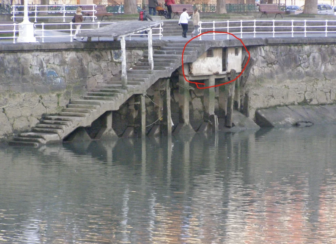 Worn stone steps leading to a dock, with several moss-covered supports. A red circle highlights a deteriorated section of the dock. Two people walk on the upper trail