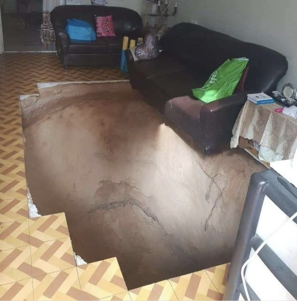 A photo showing a living room with two dark sofas and a coffee table. The floor appears to have a large, realistic-looking hole as a 3D art illusion