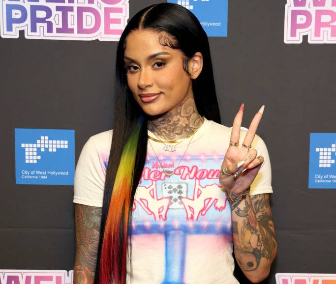 Kehlani poses at WEHO Pride event, wearing a t-shirt with a graphic design and light wash jeans, flashing a peace sign