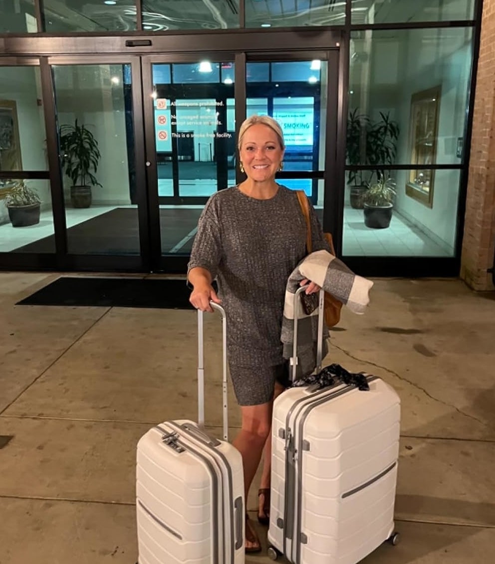 A reviewer in stylish business attire is standing outside, holding two white rolling suitcases, ready for travel