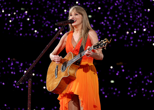 Taylor Swift performing on stage with a guitar, wearing an flowy dress, surrounded by a backdrop of lights
