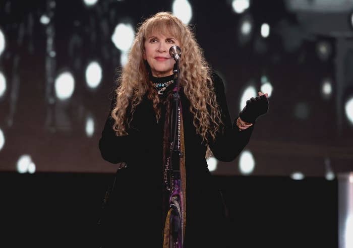 Stevie Nicks stands at a microphone on stage, wearing a dark outfit and gloves, with long, wavy hair