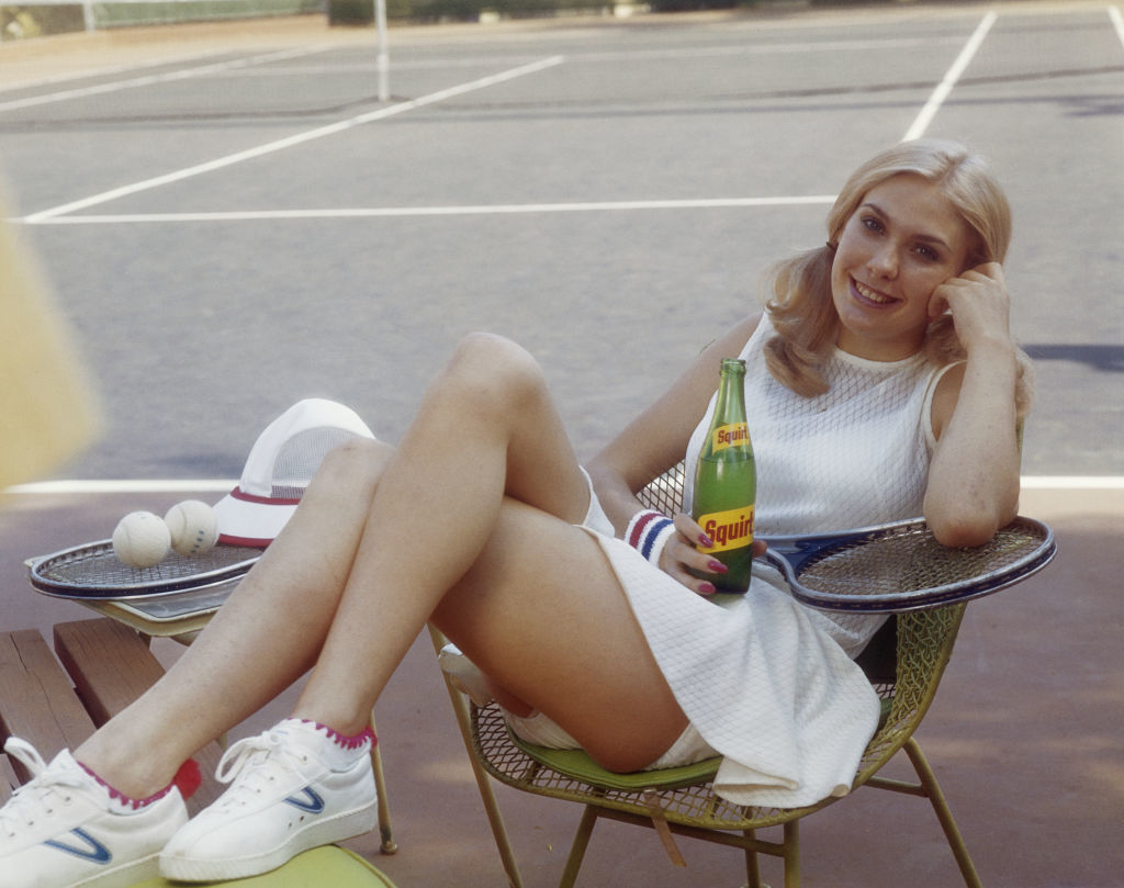 Woman in white tennis dress and sneakers sits in a chair by a tennis court, holding a Squirt soda bottle with tennis equipment nearby