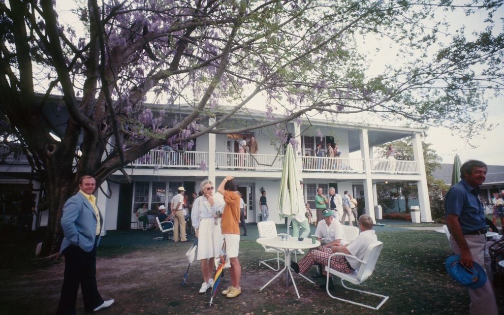 People gather in a garden setting, some sitting and others standing under a large tree in front of a two-story white building